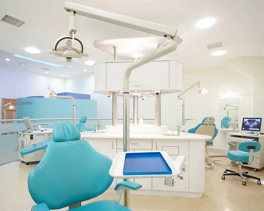 A modern orthodontic clinic featuring a turquoise dental chair with an overhead examination light and a tray for tools. The room is well-lit with white walls and sleek cabinetry. Additional dental chairs, equipment, and a computer monitor displaying a dental X-ray are visible in the background.